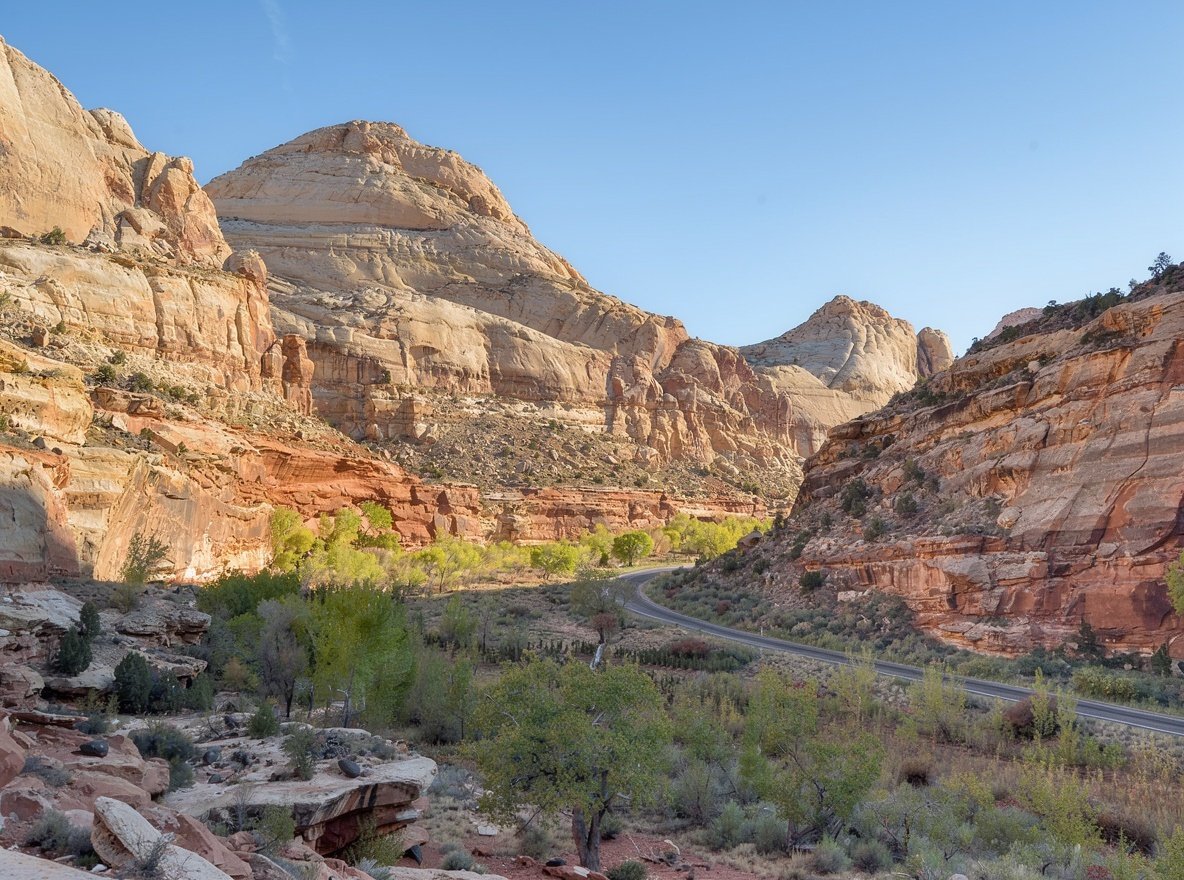 Long tilted sandstone walls in cream, grey and rust with a green valley floor below, seen from a roadside pullout in Capitol Reef